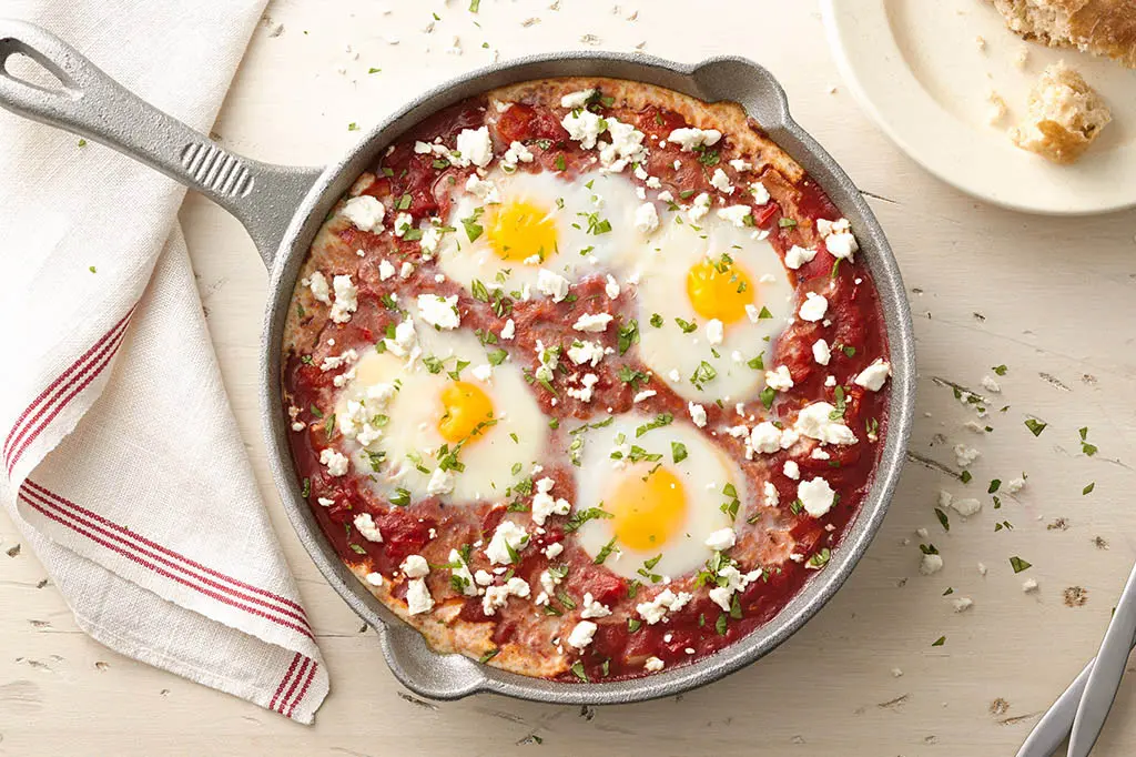 A cast iron skillet with shakshuka. Three eggs are poached in a tomato-based sauce, topped with feta cheese and parsley.
