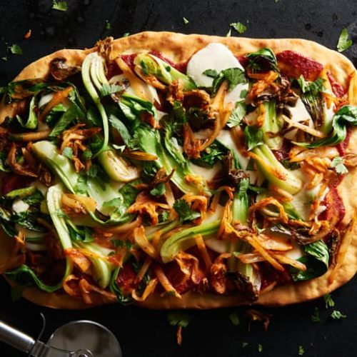 Pizza topped with tomato sauce, bok choy, spicy kimchi and herbs next to a pizza wheel
