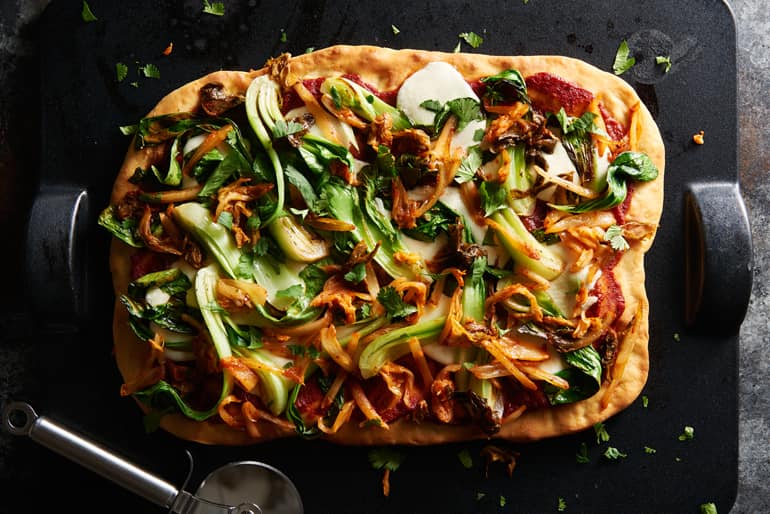 Pizza topped with tomato sauce, bok choy, spicy kimchi and herbs next to a pizza wheel