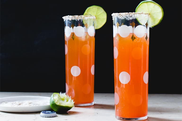 Two Muir Glen Diced Tomato water Micheladas in tall clear glasses garnished with lime and celery salt.