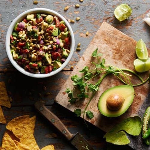 Overhead view of a bowl of avocado salsa made with Muir Glen Fire Roasted Diced Tomatoes garnished with pepitas, next to a cutting board with avocado, limes and fresh herbs.