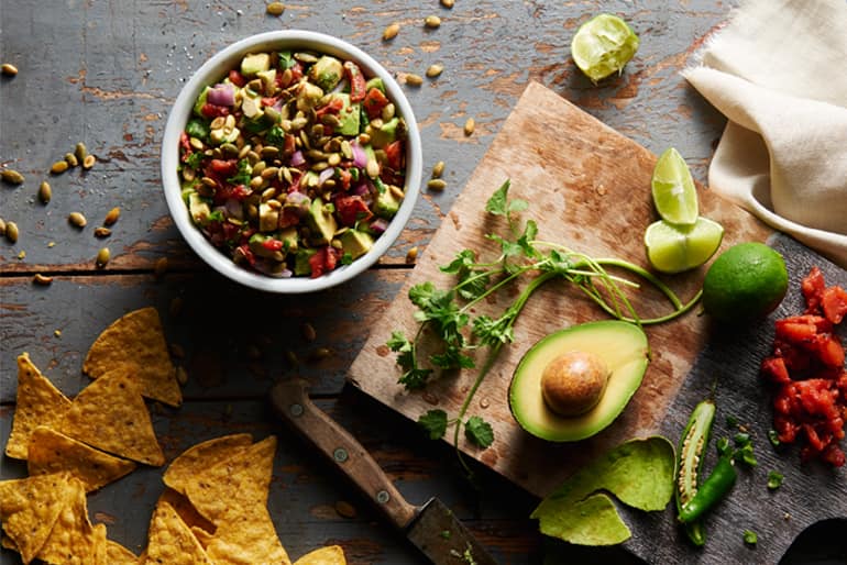 A bowl of avocado salsa garnished with pepitas, next to a cutting board with half an avocado, limes and fresh herbs