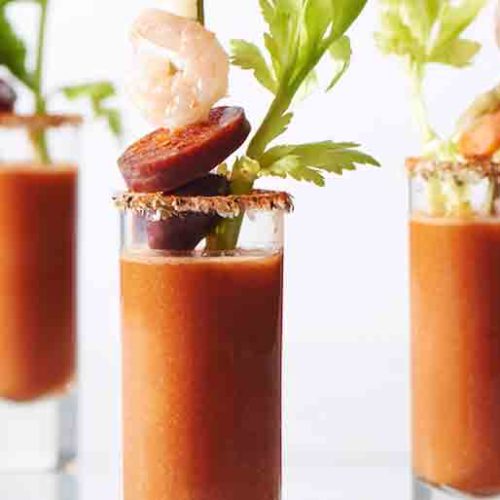 Three bloody mary tomato soup shots garnished with celery, shrimp and chorizo slices