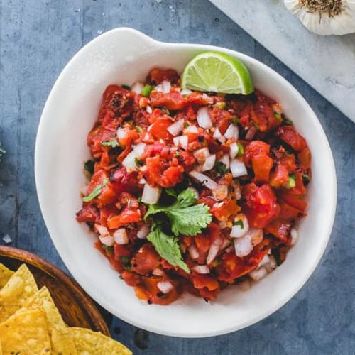 Overhead view of a bowl of salsa picada made with Muir Glen Fire Roasted Diced Tomatoes garnished with cilantro and lime served with yellow corn chips.