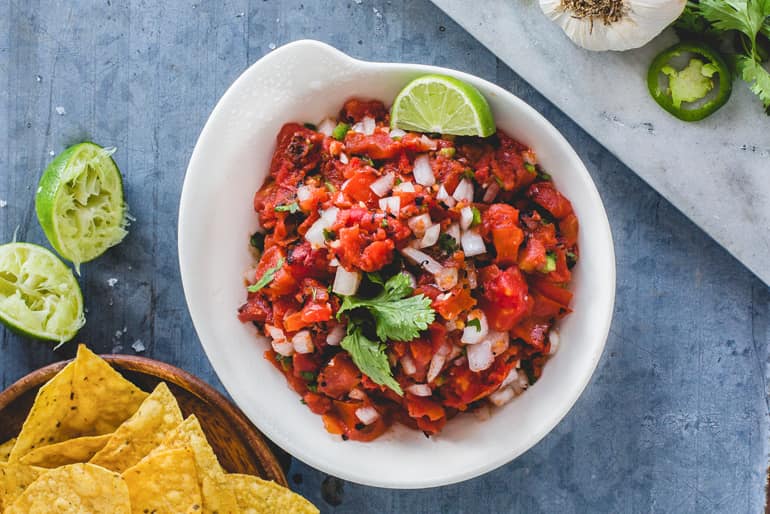 Overhead view of a bowl of salsa picada made with Muir Glen Fire Roasted Diced Tomatoes garnished with cilantro and lime served with yellow corn chips.