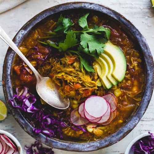 A bowl of fire roasted chipotle pozole stew garnished with avocado slices, radish, red cabbage and herbs
