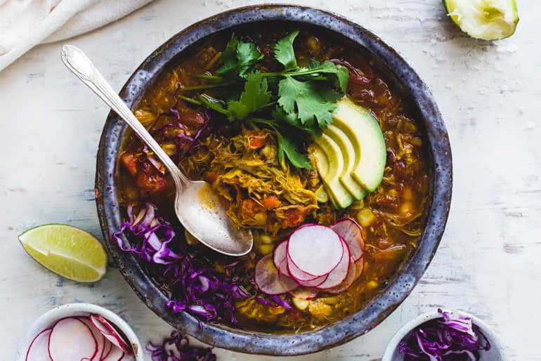 A bowl of fire roasted chipotle pozole stew garnished with avocado slices, radish, red cabbage and herbs