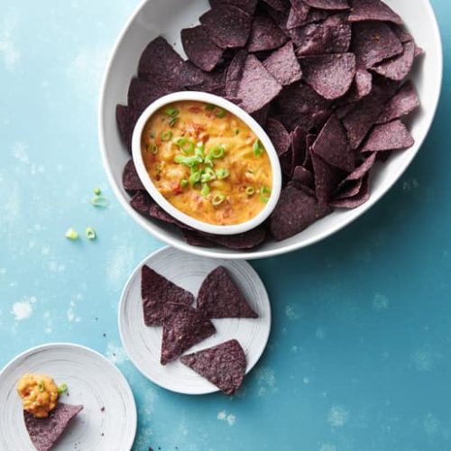 A dish full of purple tortilla chips and a smaller dish full of fire roasted tomato queso dip garnished with herbs
