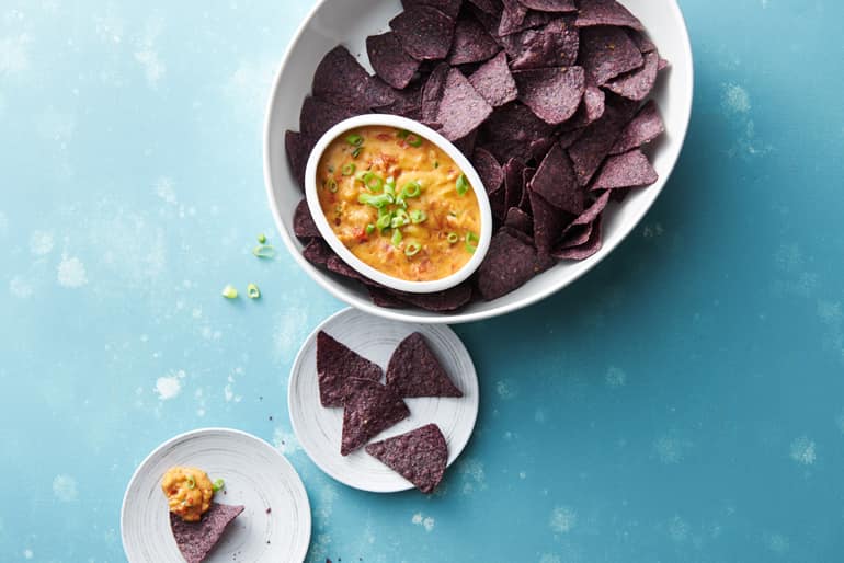 A dish full of purple tortilla chips and a smaller dish full of fire roasted tomato queso dip garnished with herbs