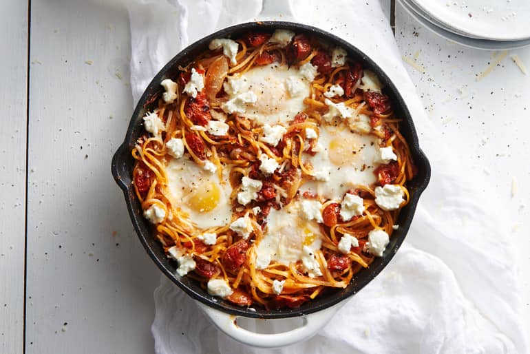 Overhead view of a skillet of spaghetti topped with Muir Glen Fire Roasted Tomato Butter Sauce, four eggs and crumbled feta cheese.