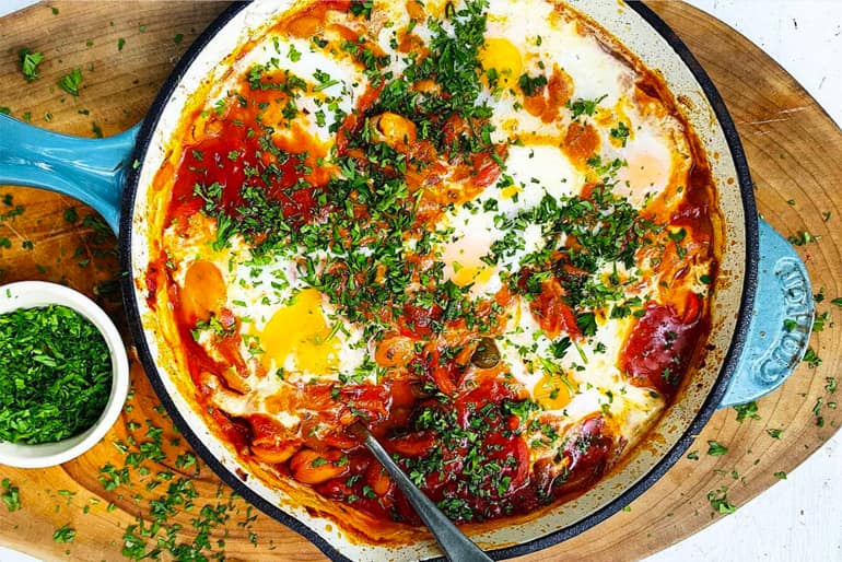 Skillet wish shakshuka topped with fresh herbs and eggs on a wooden cutting board