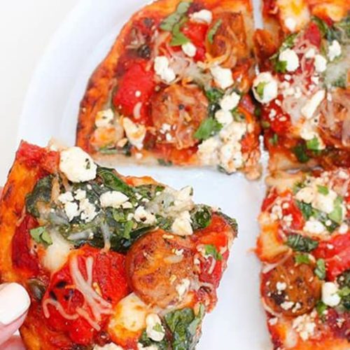 A hand removing a slice of small pizza topped with chicken sausage, onions, red peppers, leaves, and feta cheese