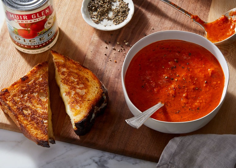 A bowl of tomato soup and a grilled cheese next to a can of Muir Glen Diced Tomatoes on a wood cutting board.