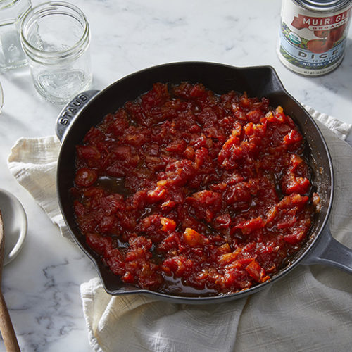 Skillet of tomato jam next to cans of Muir Glen tomatoes and a wooden spoon