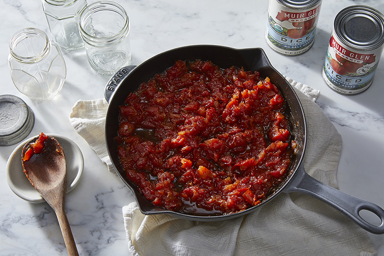 Overhead view of a skillet of tomato jam next to Muir Glen Diced Tomatoes and a wooden serving spoon on a counter.