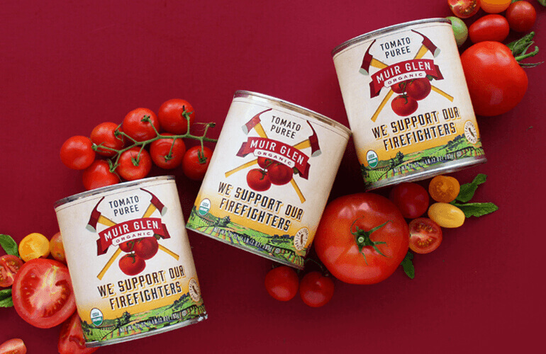 Three cans of Muir Glen tomato puree surrounded by fresh tomatoes and herbs